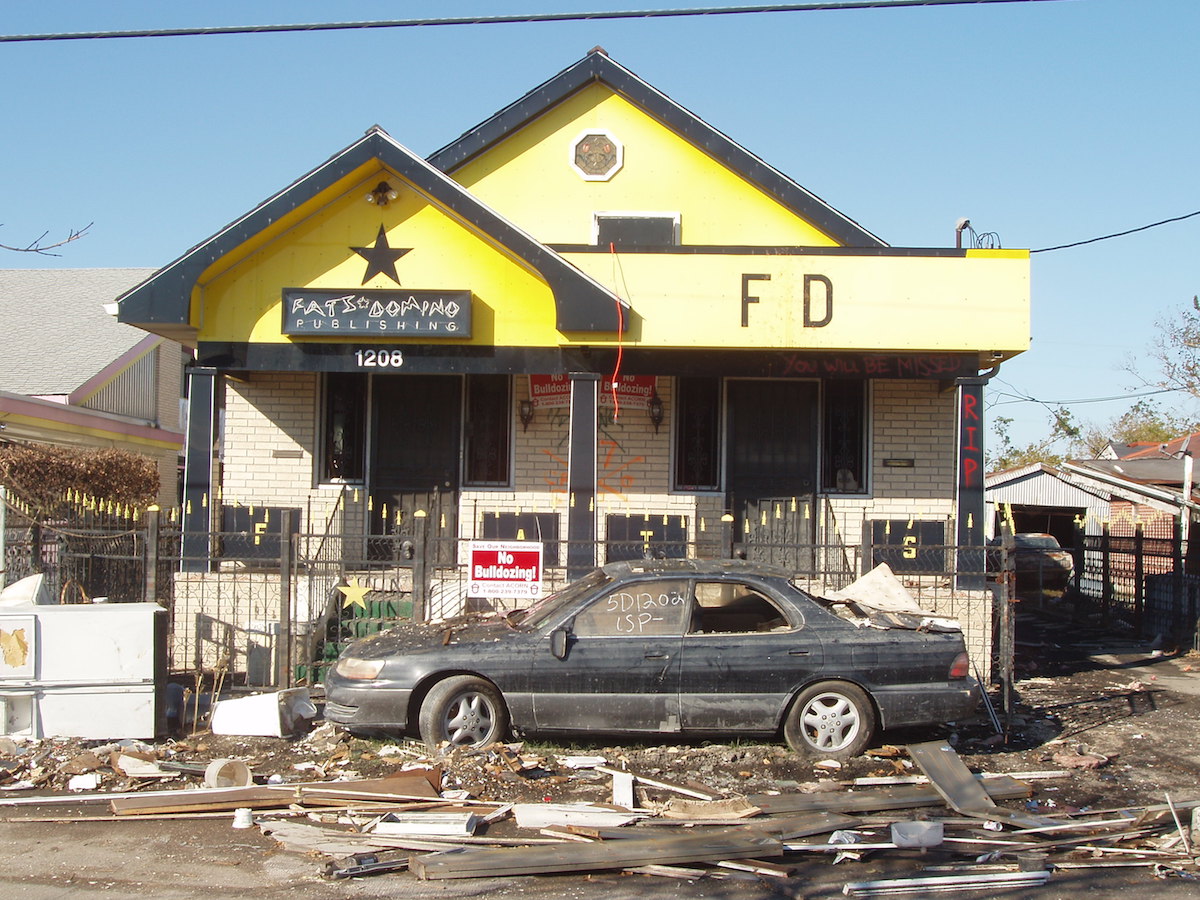 A damaged and littered, bright yellow house with a damaged, black car parked in front.