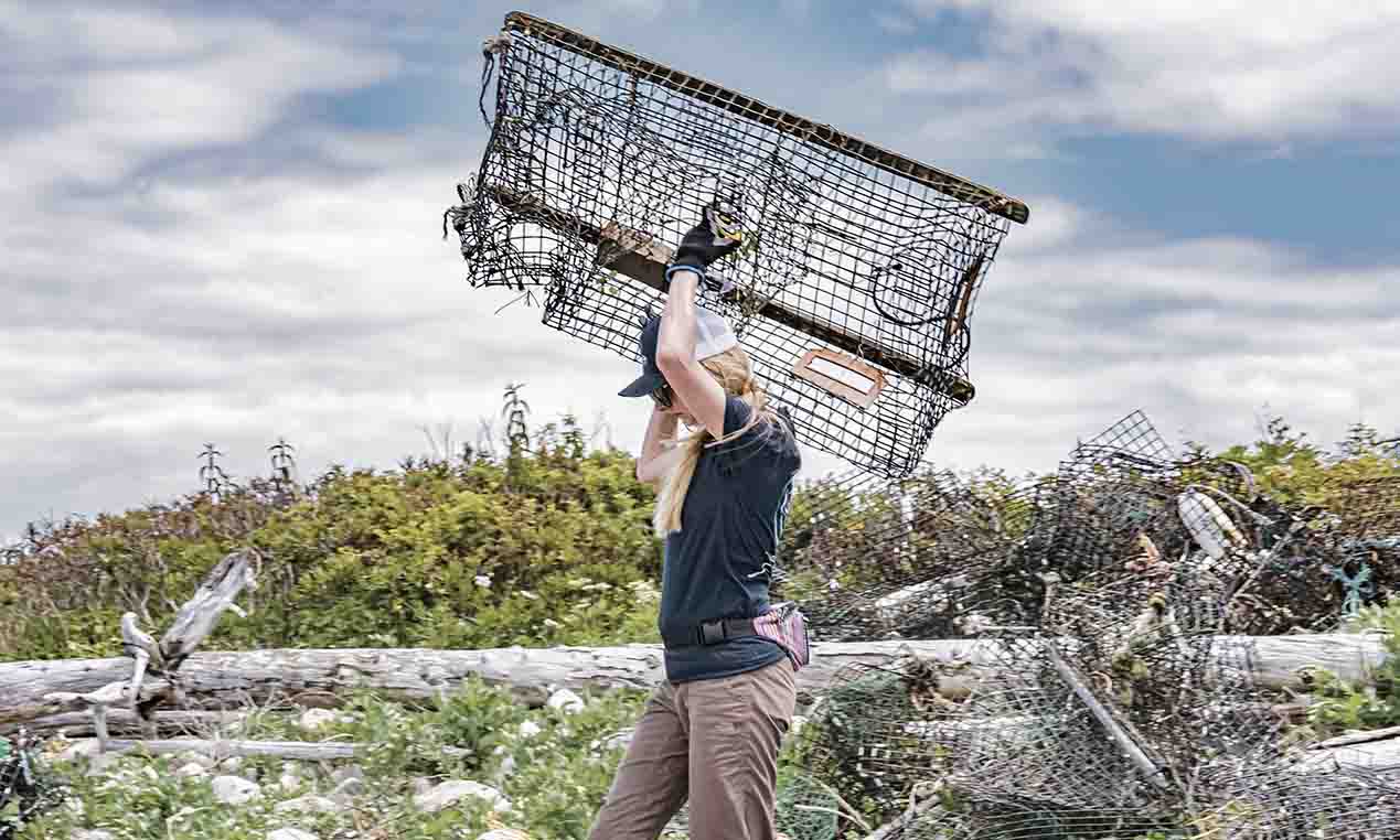 A person hauls a large lobster trap over their head as they walk across a beach.